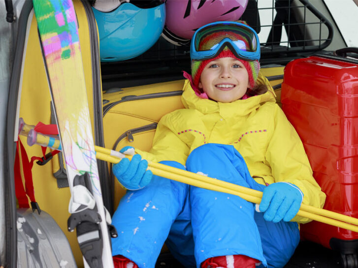 gutt i gult og blått slalomutstyr, sitter i bagasjerommet på en bil klar for påskeferie