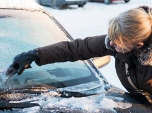 Lyshåret dame i brun parkas med pelskrage skraper frontruten på bilen.