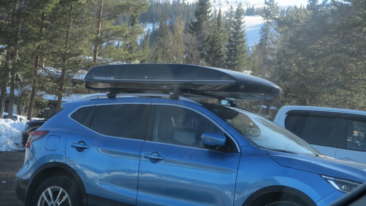 blå bil med skiboks på taket, parkert på en parkeringsplass med skog og skibakker i bakgrunnen