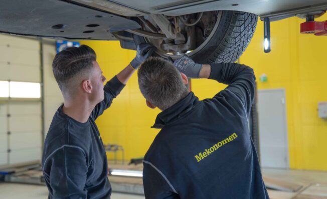 To mekanikere jobber under en bil inne på et verksted med gule vegger