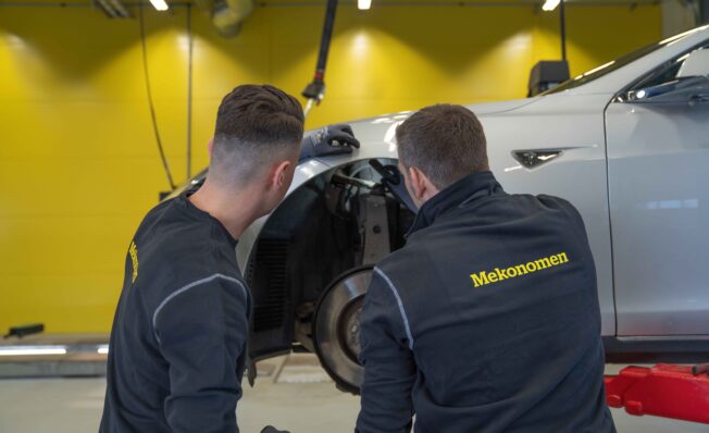 To mekanikere sjekker bremsene på hvit bil inne på mverkstede ded gule vegger