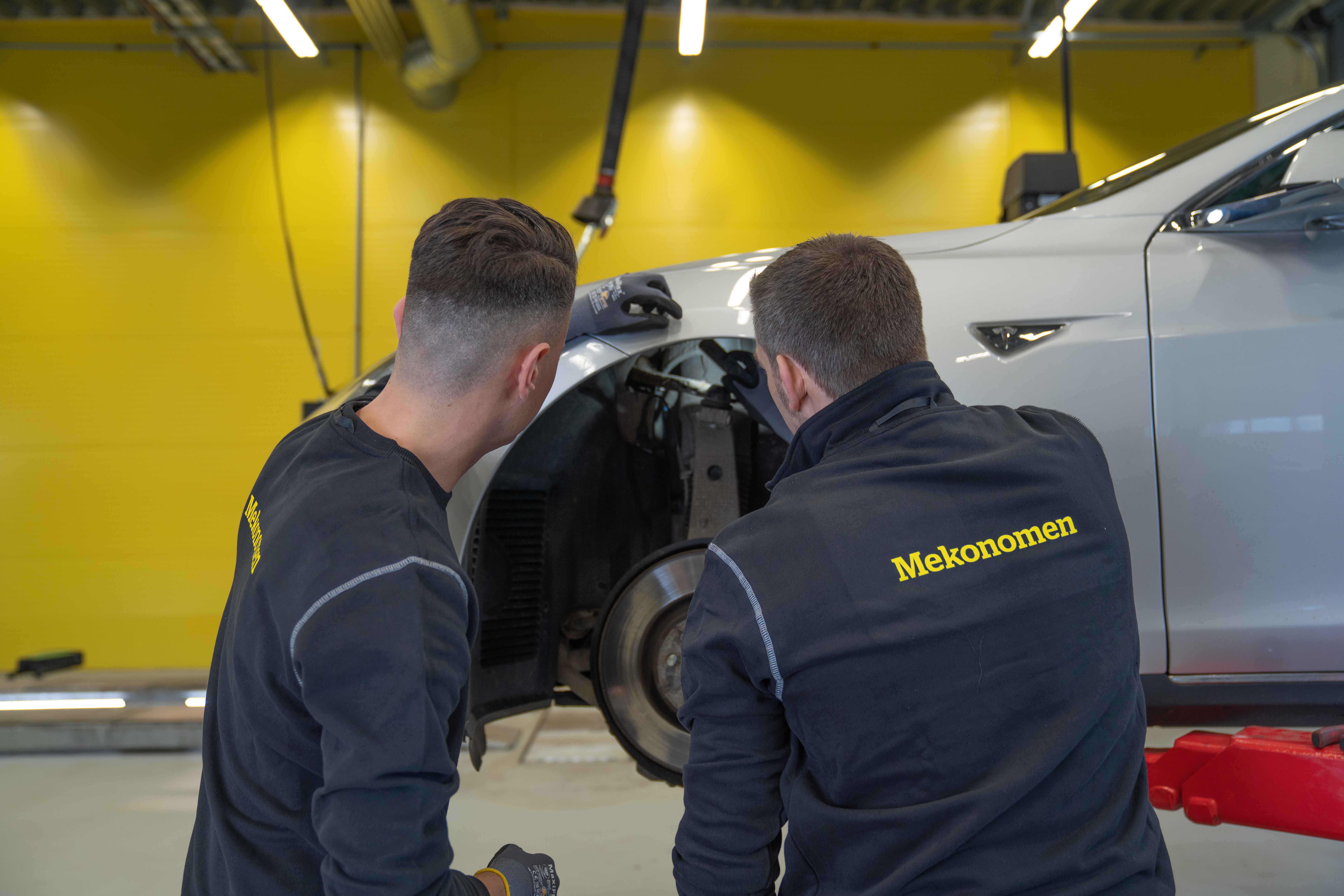 To mekanikere sjekker bremsene på hvit bil inne på mverkstede ded gule vegger