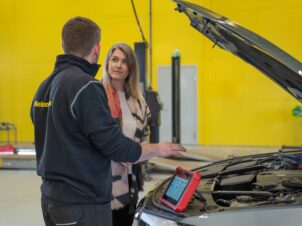 Mekaniker og kunde foran en bil inne på verkstedet med gul vegg i bakgrunnen