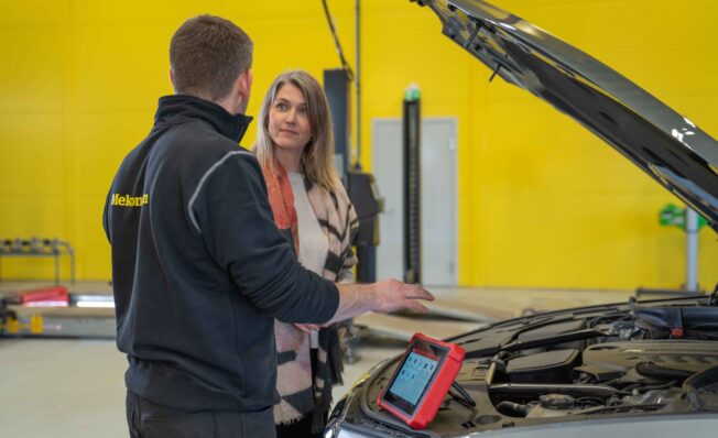 Mekaniker og kunde foran en bil inne på verkstedet med gul vegg i bakgrunnen