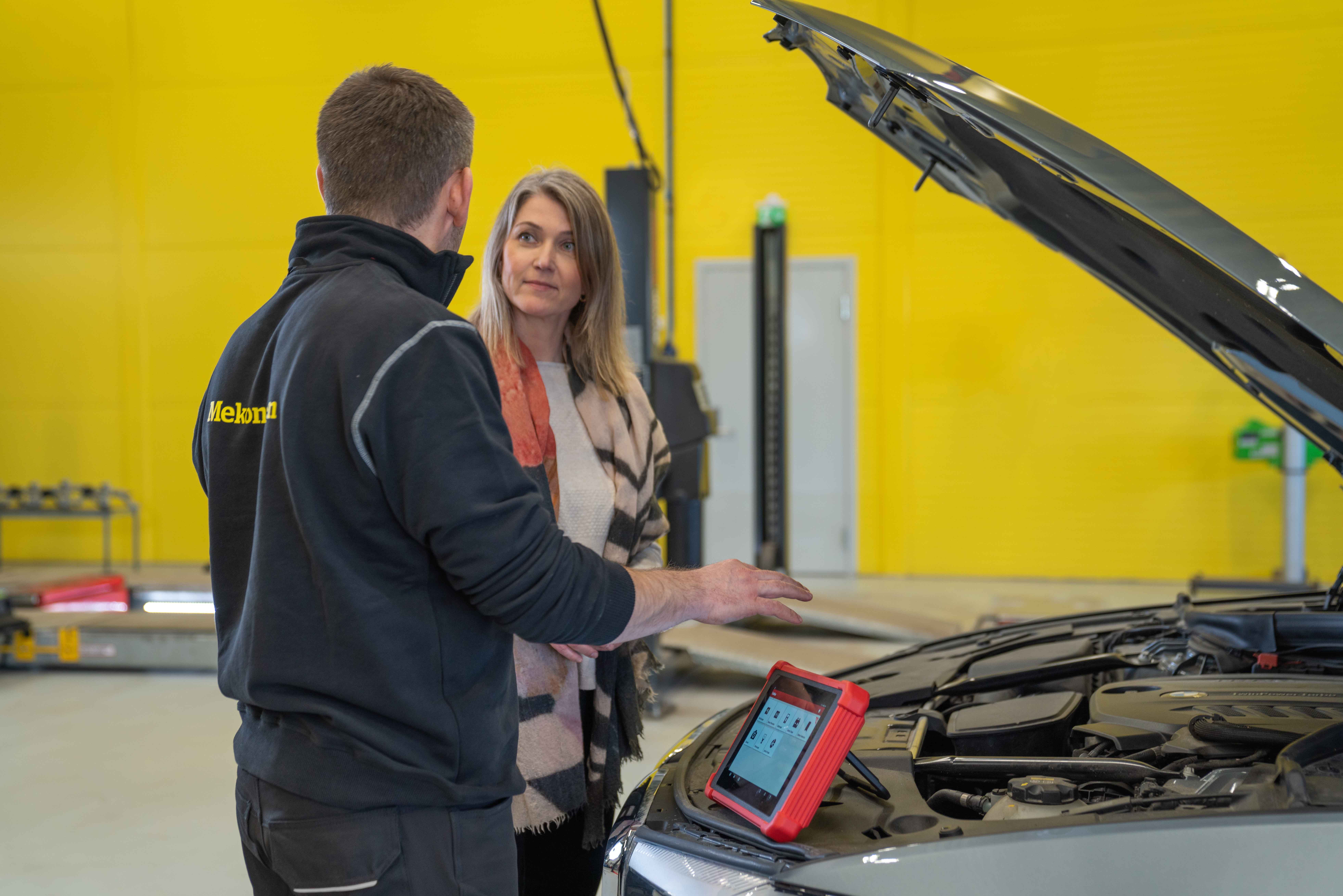 Mekaniker og kunde foran en bil inne på verkstedet med gul vegg i bakgrunnen