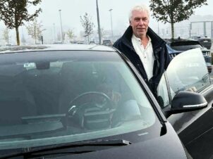 Mann står med åpen dør på sin svarte BMW I3 på en parkeringsplass en gråværsdag