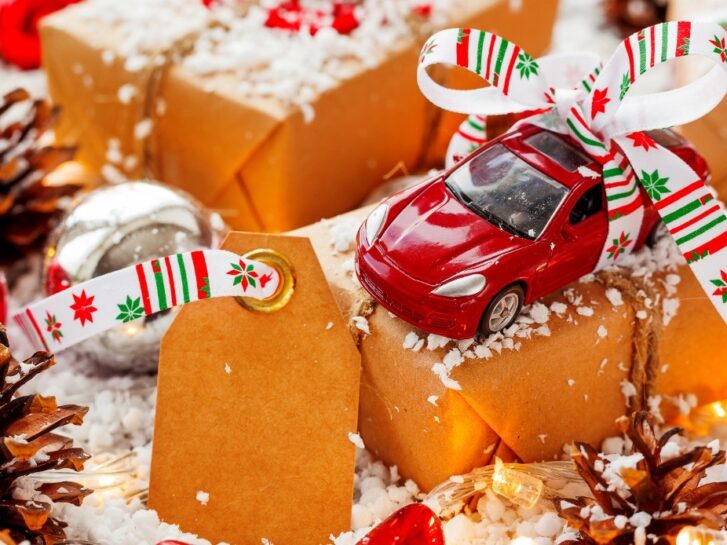 Liten rød bil med sløyfe, står oppå julegaver med oransje papir på og med kunstig snø rundt